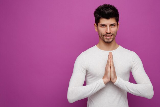 Lächelnder junger gutaussehender Mann, der in die Kamera blickt und Namaste-Geste auf violettem Hintergrund mit Kopierraum macht