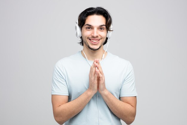 Lächelnder junger gutaussehender Mann mit Kopfhörern, der eine Namaste-Geste zeigt, während er die Kamera isoliert auf weißem Hintergrund betrachtet