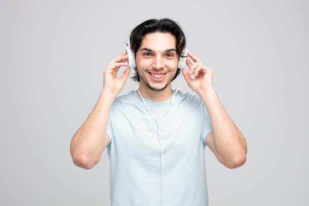 Lächelnder junger gutaussehender Mann mit Kopfhörern, der sie berührt und isoliert auf weißem Hintergrund in die Kamera schaut
