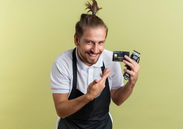 Lächelnder junger hübscher Friseur, der mit Hand auf Kreditkarte und Haarschneidemaschine lokalisiert und zeigt auf olivgrüne Wand zeigt