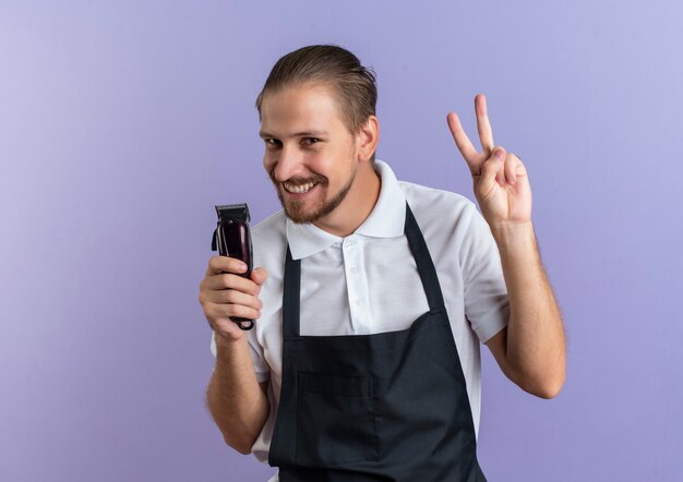 Lächelnder junger hübscher Friseur, der Uniform hält, die Haarschneidemaschinen hält und Friedenszeichen lokalisiert auf lila Wand tut