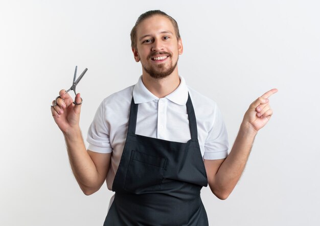 Lächelnder junger hübscher Friseur, der Uniform hält, die Schere hält und zur Seite zeigt, die auf weißer Wand lokalisiert wird