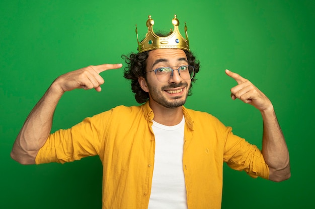 Lächelnder junger hübscher kaukasischer Mann, der Brille und Krone trägt, zeigt auf seine Krone, die Kamera lokalisiert auf grünem Hintergrund betrachtet