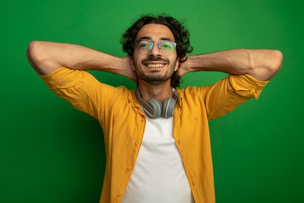 Lächelnder junger hübscher kaukasischer Mann, der eine Brille mit Kopfhörern um den Hals trägt, hält Hände hinter Kopf lokalisiert auf grüner Wand