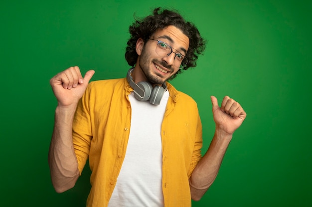 Lächelnder junger hübscher kaukasischer Mann, der eine Brille mit Kopfhörern um den Hals trägt und Kamera betrachtet, die auf sich lokalisiert auf grünem Hintergrund zeigt