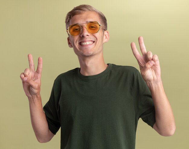 Lächelnder junger hübscher Kerl, der grünes Hemd mit Brille trägt, die Friedensgeste lokalisiert auf olivgrüner Wand zeigt