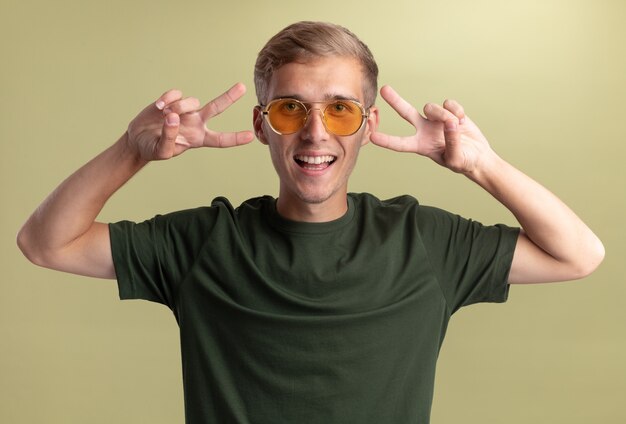Lächelnder junger hübscher Kerl, der grünes Hemd mit Brille trägt, die Friedensgeste lokalisiert auf olivgrüner Wand zeigt