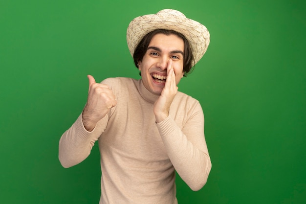 Lächelnder junger hübscher Kerl, der Hutspitzen an der Seite trägt, die Hand auf Mund lokalisiert auf grüner Wand mit Kopienraum setzen