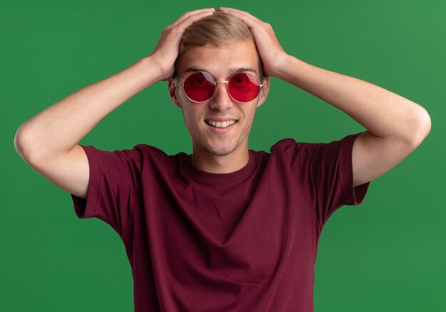 Lächelnder junger hübscher Kerl, der rotes Hemd und Brille trägt, ergriff Kopf lokalisiert auf grüner Wand