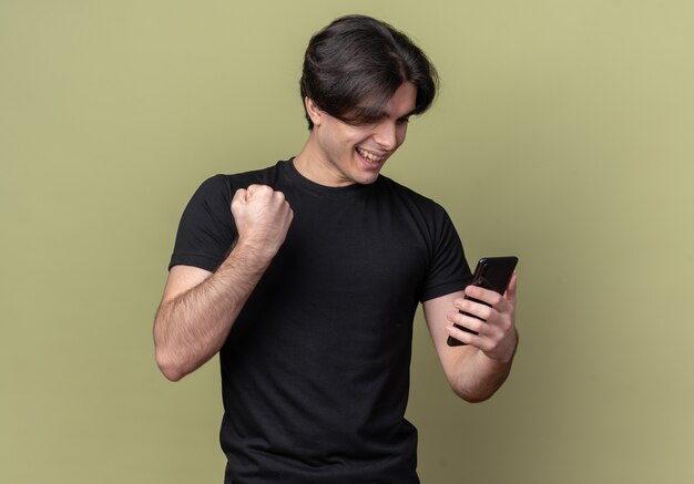 Lächelnder junger hübscher Kerl, der schwarzes T-Shirt hält Telefon hält, das ja Geste lokalisiert auf olivgrüner Wand zeigt