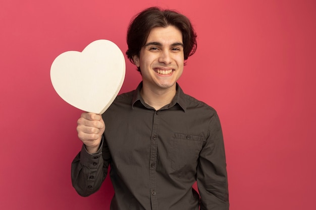 Lächelnder junger hübscher Kerl, der schwarzes T-Shirt trägt, das Herzformbox lokalisiert auf rosa Wand hält