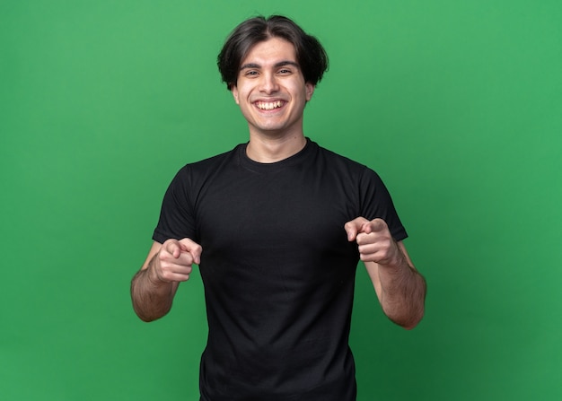 Lächelnder junger hübscher Kerl, der schwarzes T-Shirt trägt, das Sie Geste lokalisiert auf grüner Wand zeigt