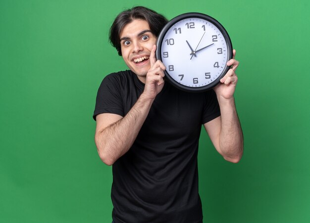 Lächelnder junger hübscher Kerl, der schwarzes T-Shirt trägt, das Wanduhr um Gesicht lokalisiert auf grüner Wand hält