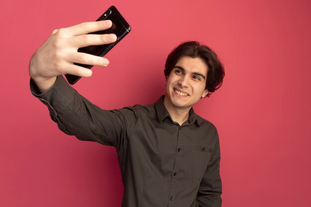 Lächelnder junger hübscher Kerl, der schwarzes T-Shirt trägt, nehmen ein Selfie lokalisiert auf rosa Wand