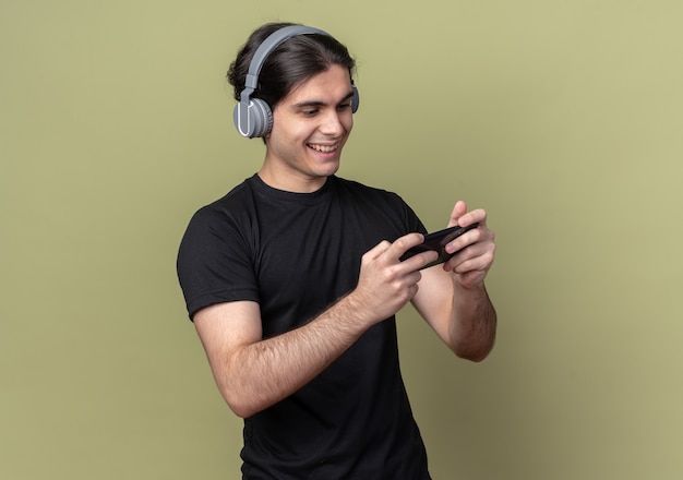 Lächelnder junger hübscher Kerl, der schwarzes T-Shirt und Kopfhörer trägt, die Spiel auf Telefon lokalisiert auf olivgrüner Wand spielen