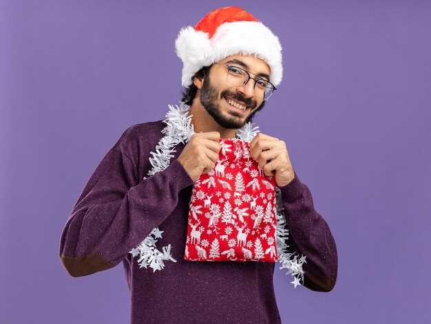 Lächelnder junger hübscher Kerl, der Weihnachtsmütze mit Girlande am Hals hält, der Weihnachtsbeutel lokalisiert auf blauem Hintergrund hält