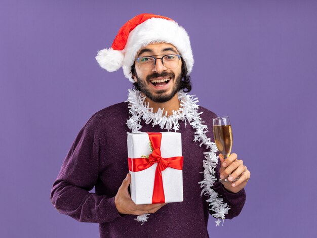 Lächelnder junger hübscher Kerl, der Weihnachtsmütze mit Girlande am Hals hält Geschenkbox mit Glas Champagner lokalisiert auf blauem Hintergrund trägt