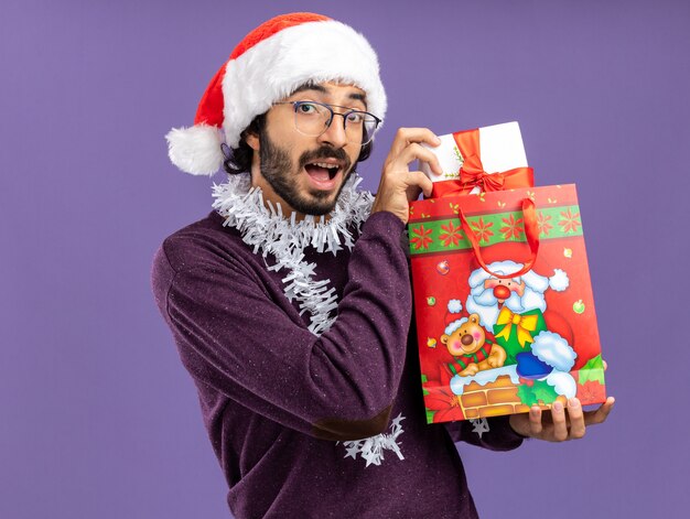 Lächelnder junger hübscher Kerl, der Weihnachtsmütze mit Girlande auf Hals hält, der Geschenktüten lokalisiert auf blauem Hintergrund trägt