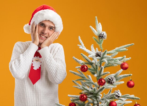 Lächelnder junger hübscher Kerl, der Weihnachtsmütze und Weihnachtsmann-Krawatte trägt, die nahe verziertem Weihnachtsbaum steht und Hände auf Gesicht hält, das Kamera lokalisiert auf orange Hintergrund betrachtet