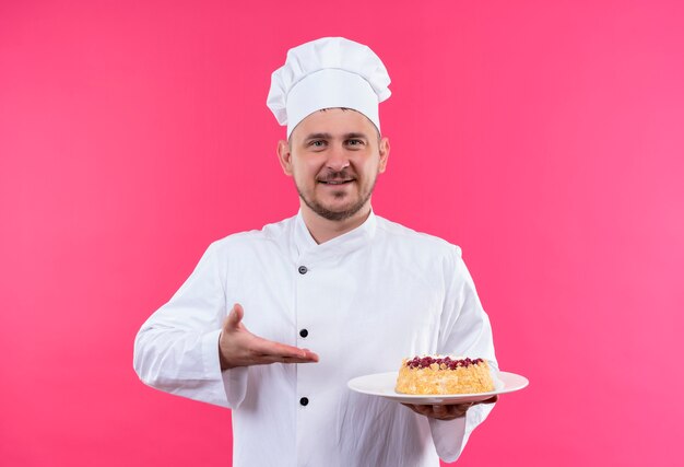 Lächelnder junger hübscher Koch in der Kochuniform, die auf Teller des Kuchens lokalisiert auf rosa Raum hält und zeigt