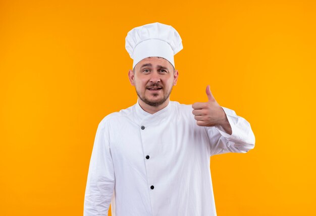 Lächelnder junger hübscher Koch in der Kochuniform, die Daumen oben auf lokalisiertem orange Raum zeigt