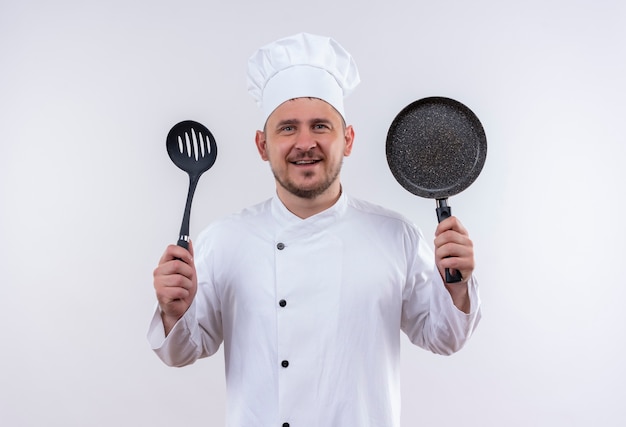 Lächelnder junger hübscher Koch in der Kochuniform, die geschlitzten Löffel und Bratpfanne auf lokalisiertem weißen Raum hält