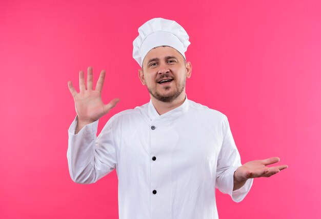 Lächelnder junger hübscher Koch in der Kochuniform, die leere Hände lokalisiert auf rosa Raum zeigt