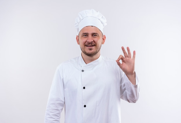 Lächelnder junger hübscher Koch in der Kochuniform, die ok Zeichen lokalisiert auf weißem Raum tut