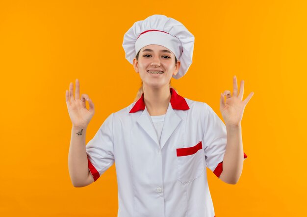 Lächelnder junger hübscher Koch in der Kochuniform mit Zahnspangen, die ok Zeichen lokalisiert auf orange Raum tun