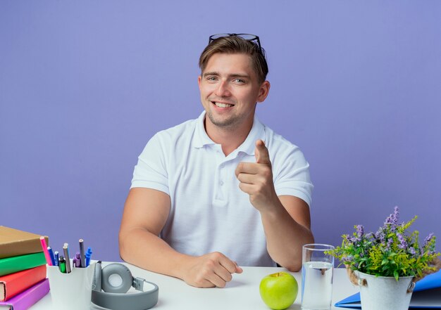 Lächelnder junger hübscher männlicher Student, der am Schreibtisch mit Schulwerkzeugen sitzt, die eine Brille auf dem Kopf tragen und Ihnen Geste zeigen, die auf Blau lokalisiert wird