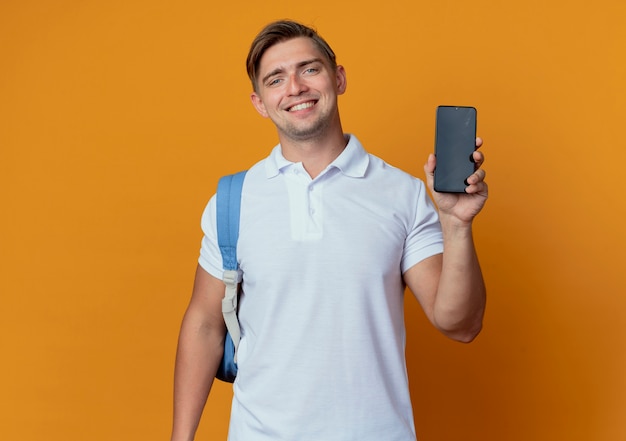 Lächelnder junger hübscher männlicher Student, der Rückentasche hält Telefon lokalisiert auf Orange