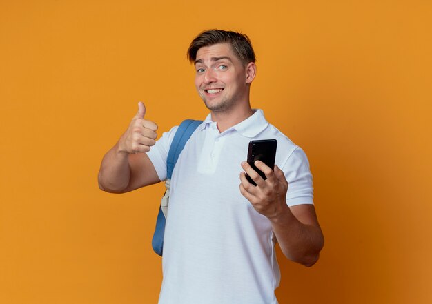 Lächelnder junger hübscher männlicher Student, der Rückentasche hält Telefon und seinen Daumen oben auf Orange isoliert trägt