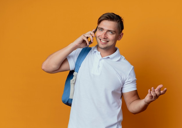 Lächelnder junger hübscher männlicher Student, der Rückentasche trägt, spricht am Telefon