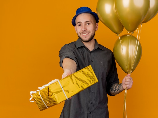 Lächelnder junger hübscher slawischer party-typ, der partyhut hält, der ballons hält, die geschenkverpackung in richtung kamera lokalisiert auf orange hintergrund strecken