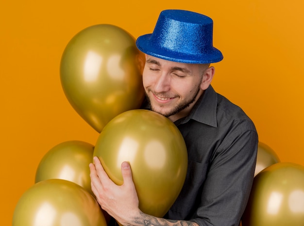 Kostenloses Foto lächelnder junger hübscher slawischer party-typ, der partyhut trägt, der unter luftballons steht, die einen mit geschlossenen augen lokalisiert auf orangeem hintergrund ergreifen