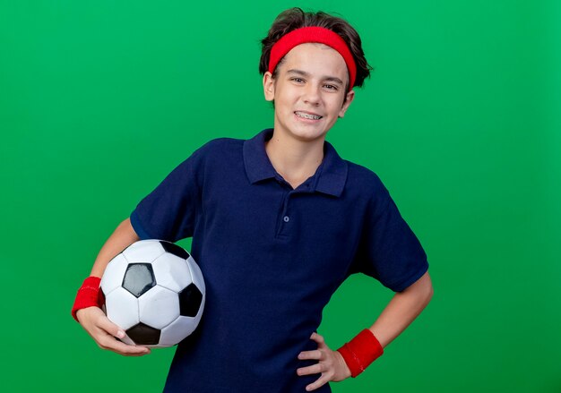 Lächelnder junger hübscher sportlicher Junge, der Stirnband und Armbänder mit Zahnspangen hält, die Fußball halten Hand auf Taille halten, die Kamera lokalisiert auf grünem Hintergrund mit Kopienraum hält