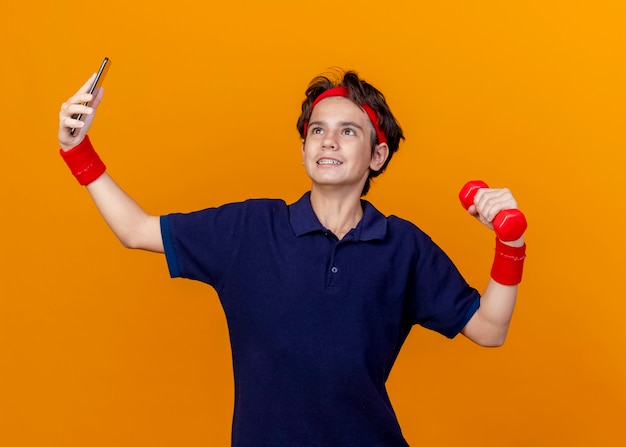 Lächelnder junger hübscher sportlicher Junge, der Stirnband und Armbänder mit Zahnspangen hält, die Hantel halten, die nach oben genommenes Selfie lokalisiert auf orange Wand hält