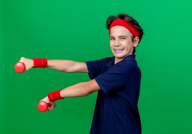 Lächelnder junger hübscher sportlicher Junge, der Stirnband und Armbänder mit Zahnspangen trägt, die in der Profilansicht stehen, die Hanteln ausdehnt, die Kamera lokalisiert auf grünem Hintergrund mit Kopienraum betrachten