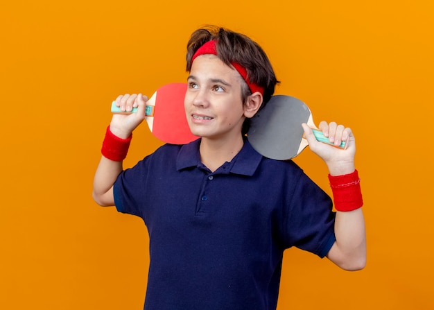 Lächelnder junger hübscher sportlicher Junge, der Stirnband und Armbänder mit Zahnspangen trägt, die Tischtennisschläger auf Schultern halten, die Seite lokalisiert auf orange Hintergrund betrachten