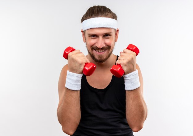 Lächelnder junger hübscher sportlicher Mann, der Stirnband und Armbänder hält, die Hanteln halten, die auf weißem Raum isoliert suchen