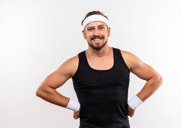 Lächelnder junger hübscher sportlicher Mann, der Stirnband und Armbänder trägt, die Hände auf Taille lokalisiert auf weißem Raum setzen