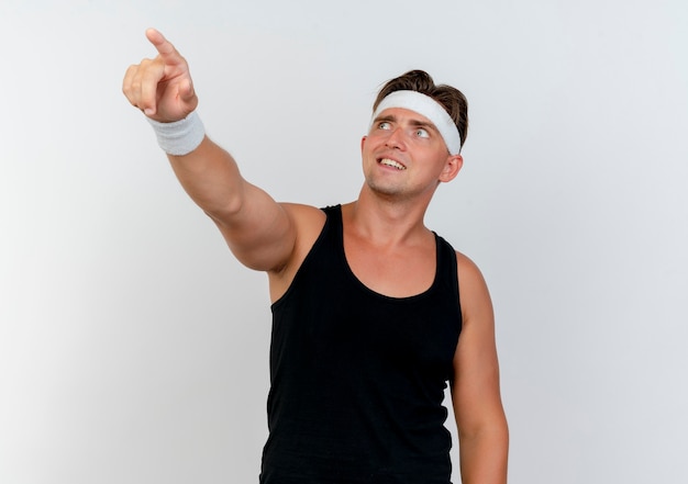 Lächelnder junger hübscher sportlicher Mann, der Stirnband und Armbänder trägt, die oben schauen und auf Seite zeigen, die auf weißer Wand lokalisiert ist