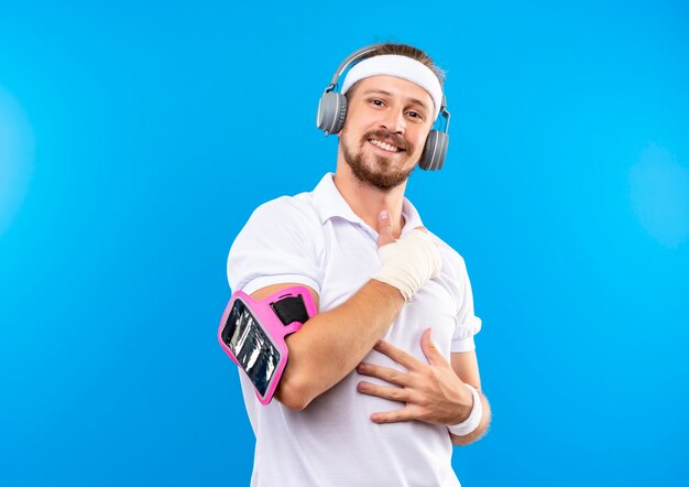 Lächelnder junger hübscher sportlicher Mann, der Stirnband und Armbänder und Kopfhörer mit Telefonarmband trägt, die Hände auf Bauch und Brust mit verletztem Handgelenk setzen, das mit Verband umwickelt wird, der auf blauem Raum isoliert wird