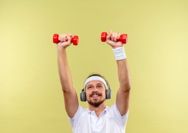 Lächelnder junger hübscher sportlicher Mann, der Stirnband und Armbänder und Kopfhörer trägt, die Hanteln heben, die lokal auf Grünfläche suchen