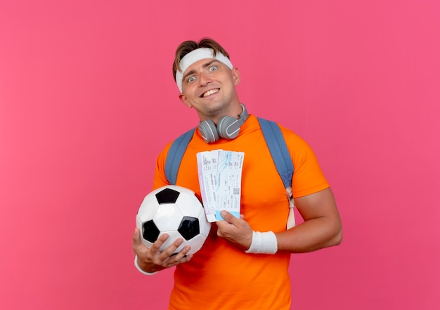 Lächelnder junger hübscher sportlicher Mann, der Stirnband und Armbänder und Rückentasche mit Kopfhörern am Hals trägt, die Flugtickets und Fußball lokalisiert auf rosa Wand halten