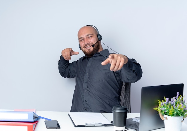 Lächelnder junger kahler Callcenter-Mann, der Headset trägt, das am Schreibtisch mit Arbeitswerkzeugen sitzt, die Sie Geste an der Front lokalisiert auf weißer Wand tun