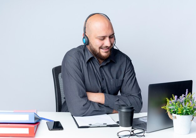 Lächelnder junger kahler Callcenter-Mann, der Headset trägt, das mit geschlossener Haltung am Schreibtisch mit Arbeitswerkzeugen sitzt, die Laptop lokalisiert auf weißer Wand betrachten