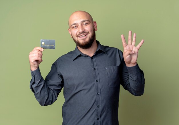 Lächelnder junger kahler Callcenter-Mann, der Kreditkarte hält, die vier mit der Hand lokalisiert auf olivgrüner Wand zeigt