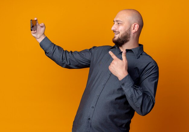 Lächelnder junger kahlköpfiger Callcenter-Mann, der selfie nimmt und auf Handy zeigt, das auf orange Wand lokalisiert ist