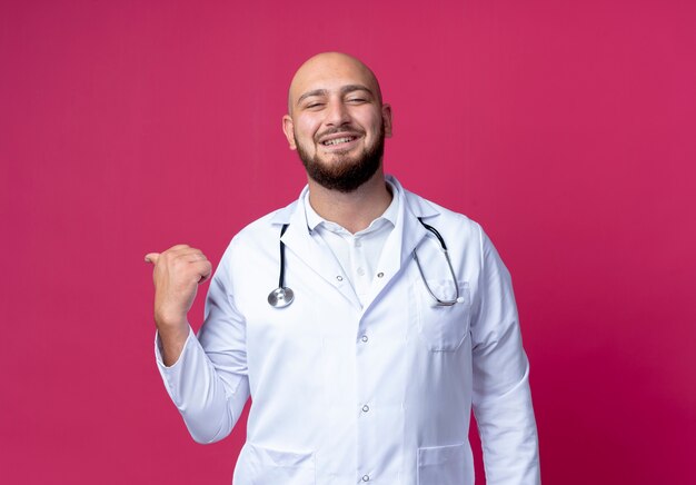 Lächelnder junger kahlköpfiger männlicher Arzt, der medizinische Robe und Stethoskoppunkte an der Seite trägt, lokalisiert auf rosa mit Kopienraum